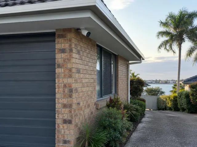 White CCTV camera mounted under a roof eave on a brick home exterior with driveway and water view in Runaway Bay